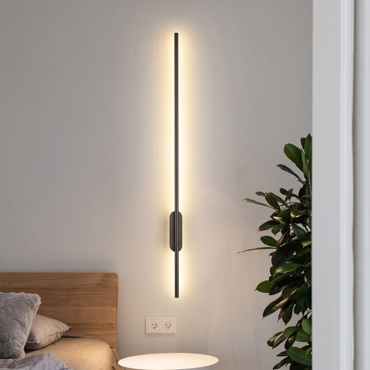 A long black minimalist wall sconce mounted beside a wooden bed frame, casting a warm ambient light on the wall.